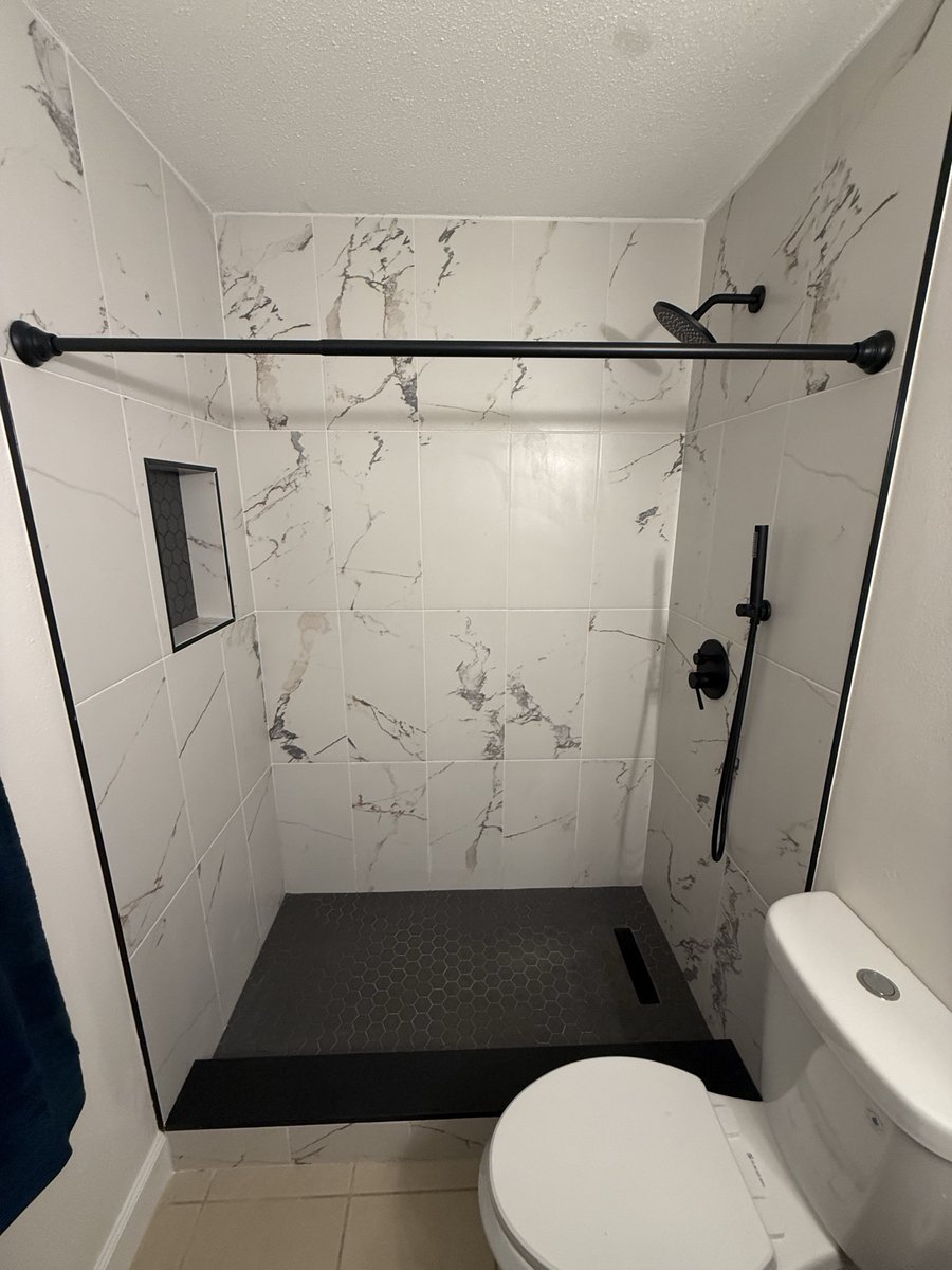 Walk-in shower by KM — marble-look tile, black fixtures, and dark hex tile floor
